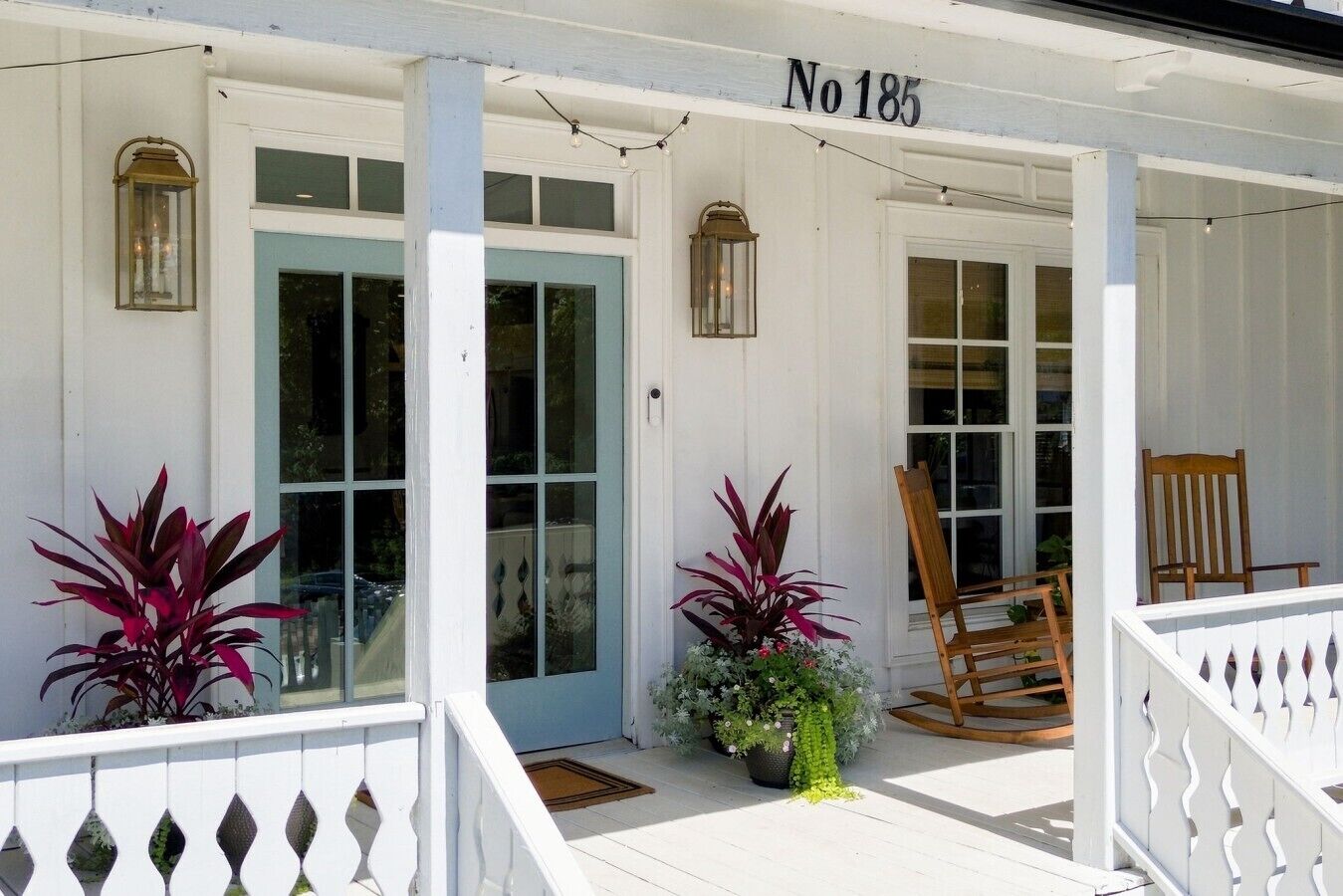 Un bâtiment blanc avec une porte bleue, un fauteuil à bascule en bois et des plantes en pot.