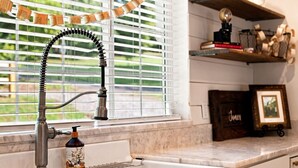A kitchen with a sink, a window with blinds, a shelf with books and decorative items, and a bottle of soap.