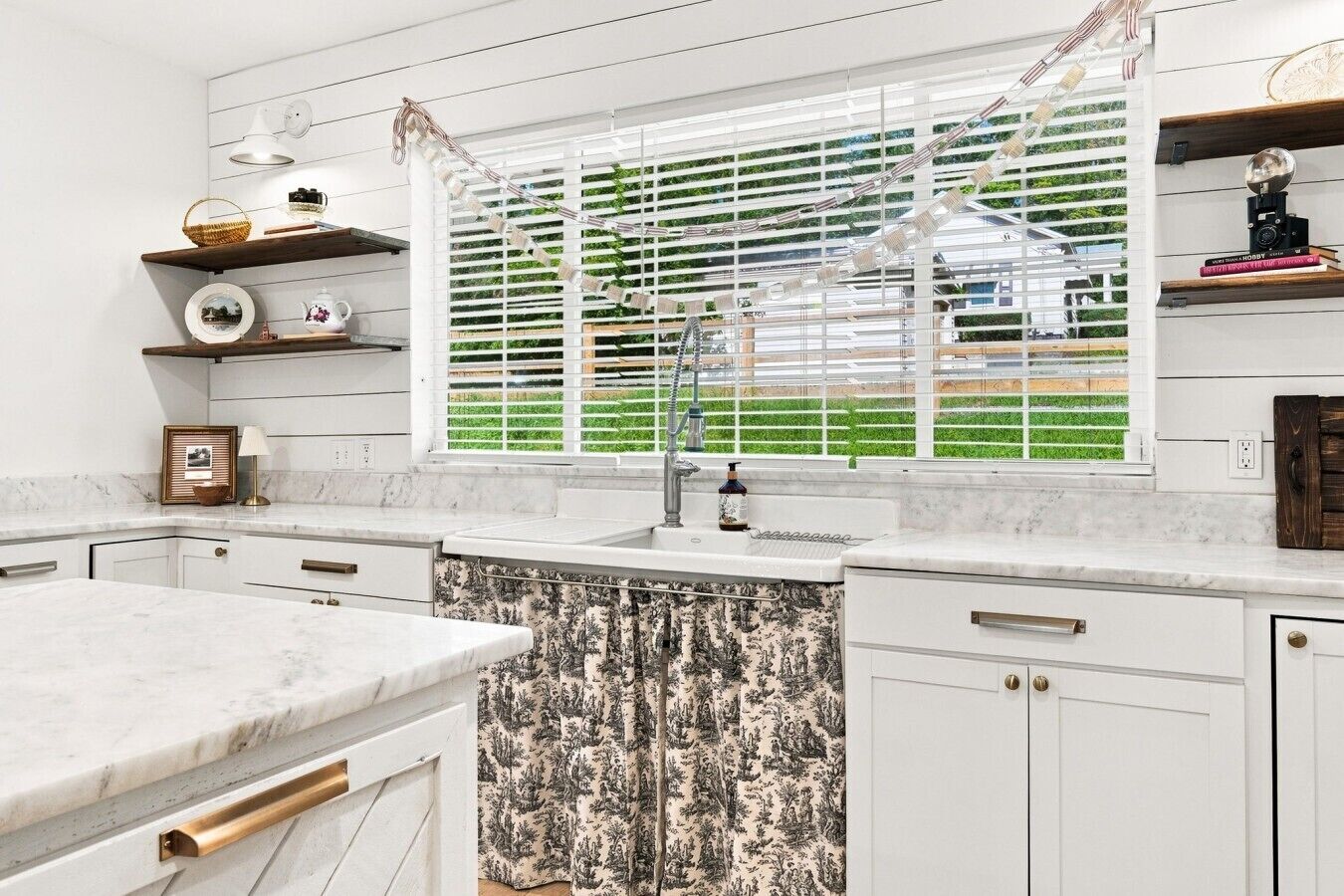 A kitchen with white cabinetry, a marble countertop, a window with blinds, and a view of greenery outside.