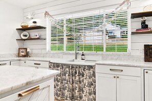 Cocina con armarios blancos, encimera de mármol, ventana con persianas y vistas al exterior con vegetación.