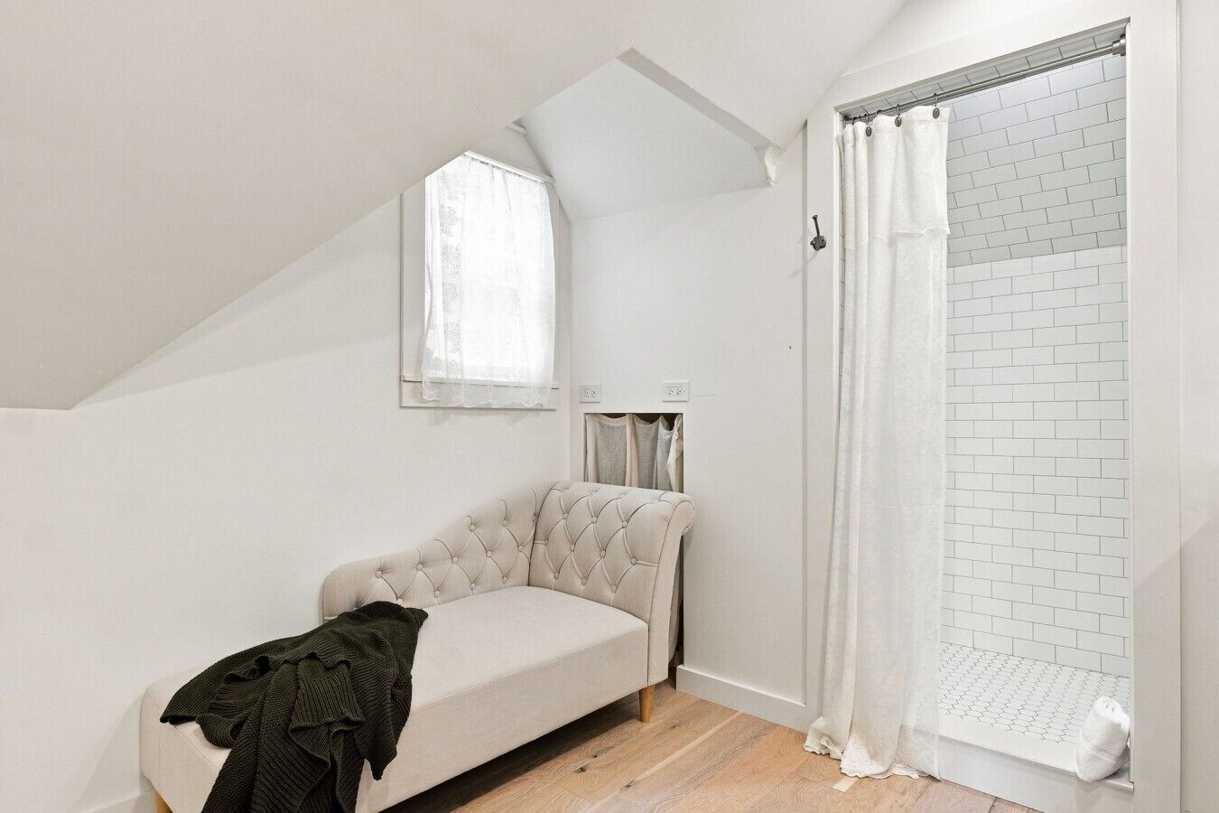A small, white-walled room with a sloped ceiling, a single window with a sheer curtain, a white cushioned bench, and a white shower area with a tiled wall.