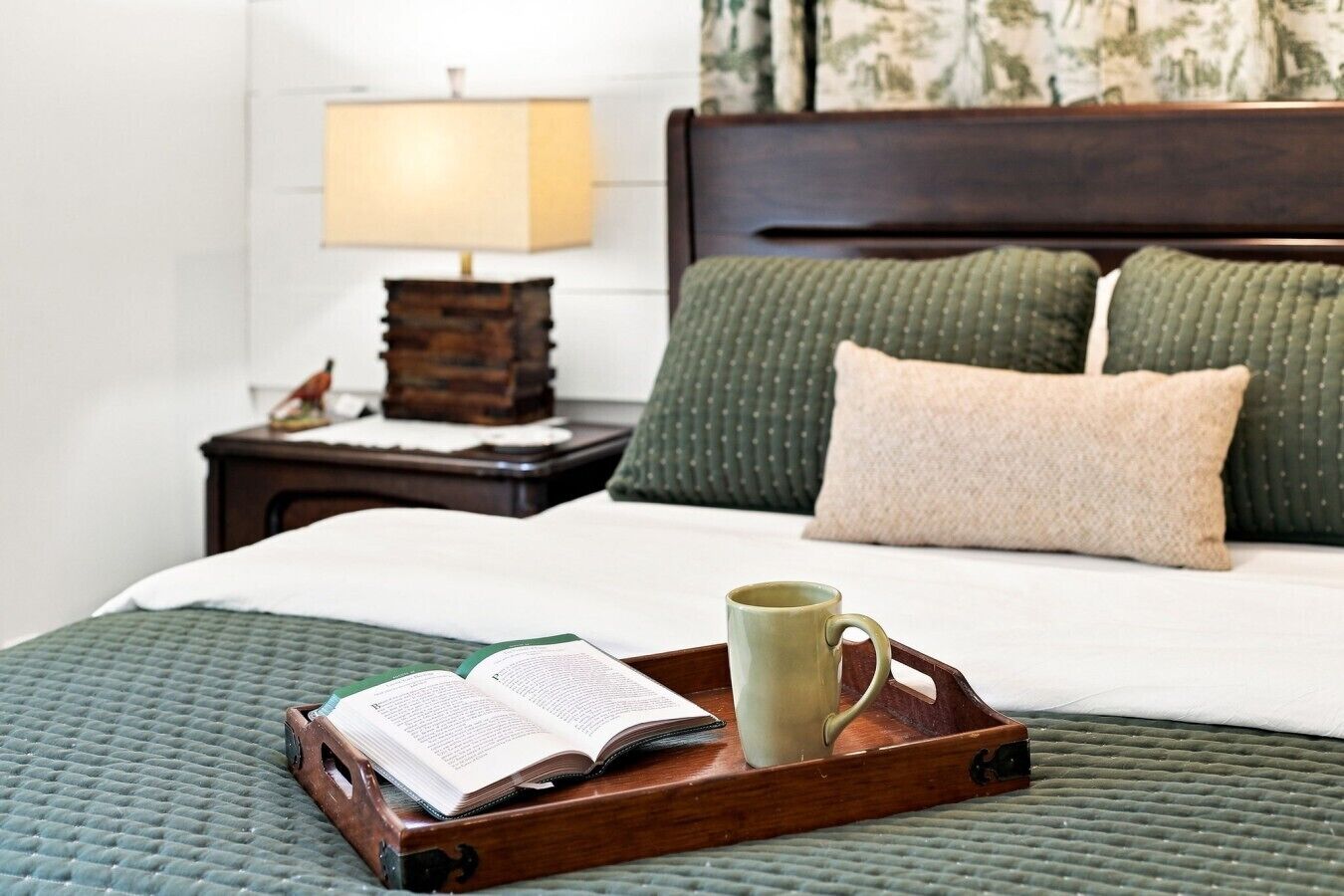 A neatly made bed with a green quilt, a wooden tray with a book and mug, a nightstand with a lamp, and a patterned wall.