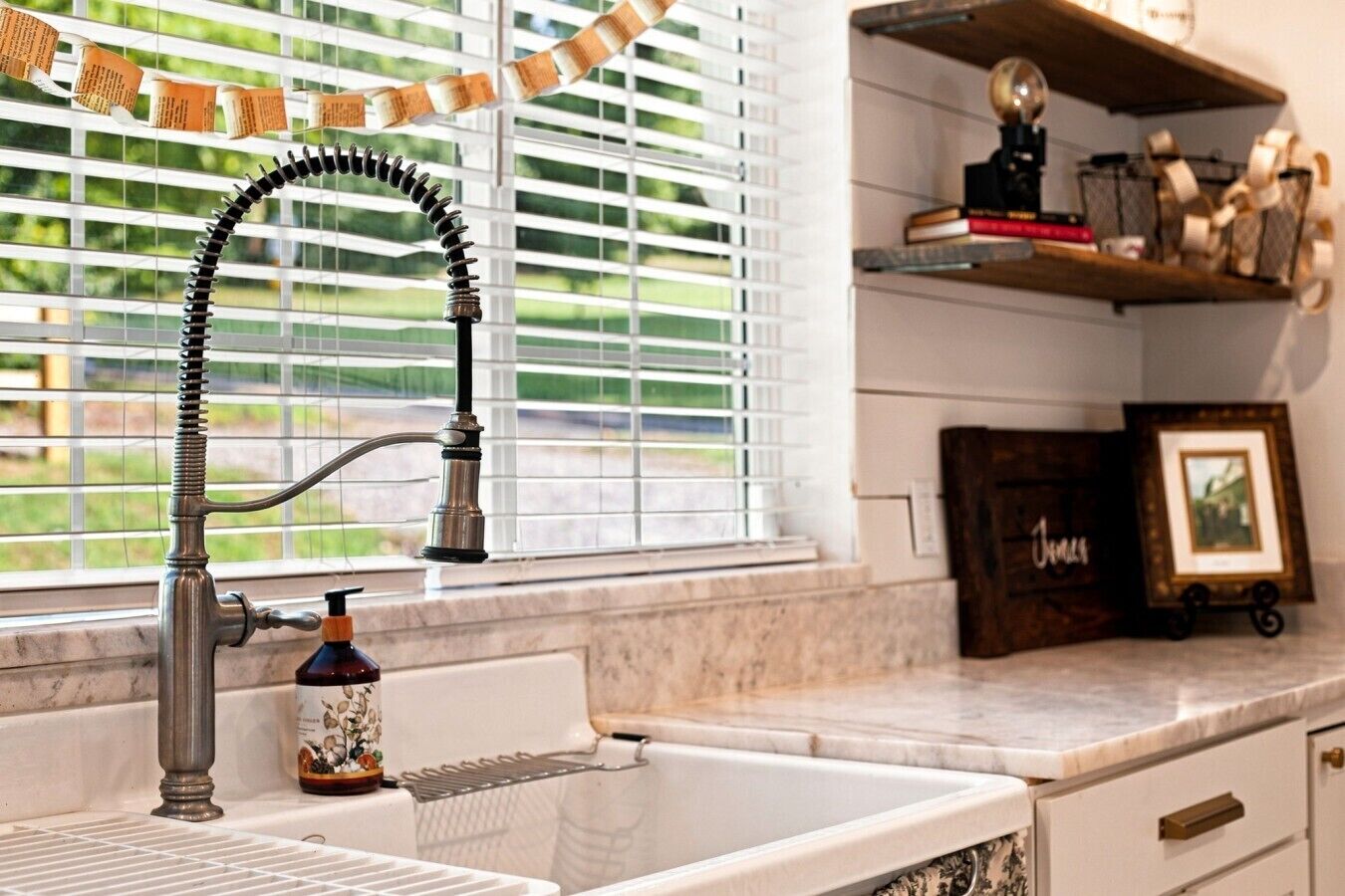 A kitchen with a sink, a window with blinds, a shelf with books and decorative items, and a bottle of soap.