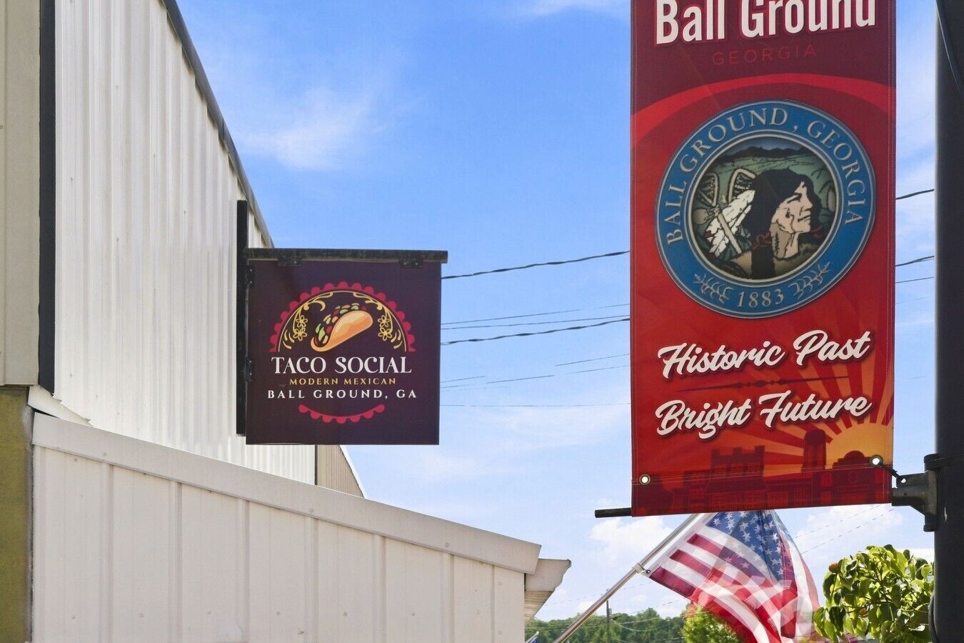 Deux panneaux, l’un indiquant « Taco Social » et l’autre « Ball Ground, Géorgie », avec un drapeau américain en arrière-plan.