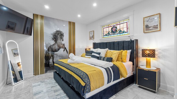 Ein modernes Schlafzimmer mit einem großen Bett, einem Nachttischchen mit Lampe, einem Spiegel sowie einer Wand mit einem Gemälde von einem Pferd und einem Buntglasfenster.