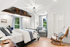 A bedroom with a bed, a rocking chair, a sofa, a wooden shelf, and a view of greenery outside.