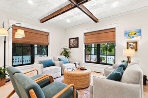 A spacious living area with a high ceiling, large windows with blinds, and a central round coffee table.