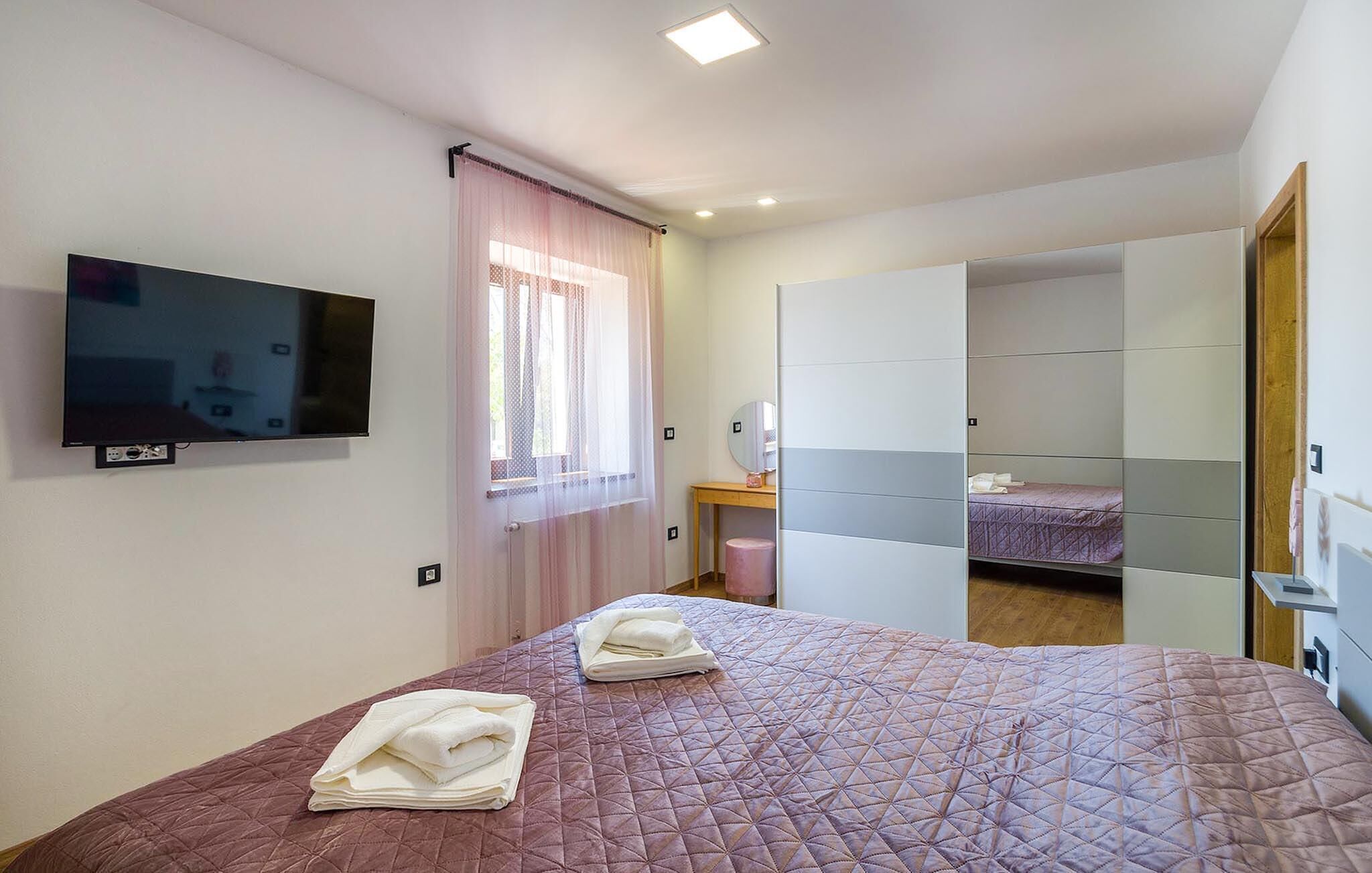 A bedroom with a bed, a TV mounted on the wall, a window with curtains, a bedside table, and a wardrobe.
