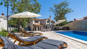 A poolside area with lounge chairs and umbrellas, a stone pool, and a building with a tiled roof. - Stunning home in Sajini (Sajini)