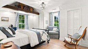 Ein Schlafzimmer mit einem Bett, einem Schaukelstuhl, einer Couch, einem Holzregal und Blick ins Grüne.