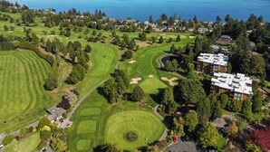 An aerial view of a golf course with a clubhouse, surrounded by residential buildings and a large body of water. - Cordova Bay Living (Victoria)