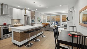 A modern kitchen with a central island, stainless steel appliances, and a dining area with a dark wooden table and bar stools. - Cordova Bay Living (Victoria)