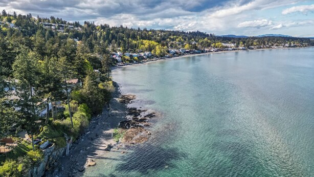 A coastal area with a clear view of a beach, rocky shoreline, and a forested hillside. - Cordova Bay Living (Victoria)