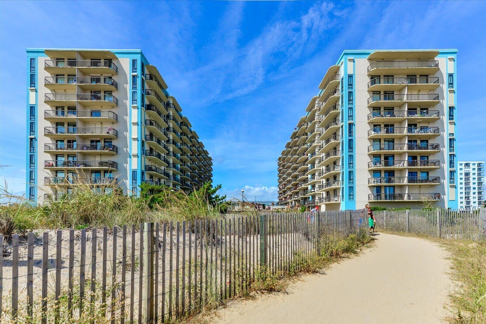 Un hôtel situé en bord de mer, doté de nombreux balcons, d’un sentier sablonneux menant à la plage et d’une clôture en bois.