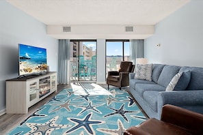 A modern hotel room with a balcony, a large flat-screen TV, a sofa, and a starfish-patterned rug. - Two Bedroom Beachfront Condo with Great Amenities (Ocean City)