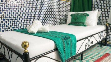 Una cama con colchón blanco, manta verde y almohadas blancas. El cabecero es negro con detalles dorados. La habitación tiene baldosas azules y blancas con diseño en las paredes y un suelo a cuadros.