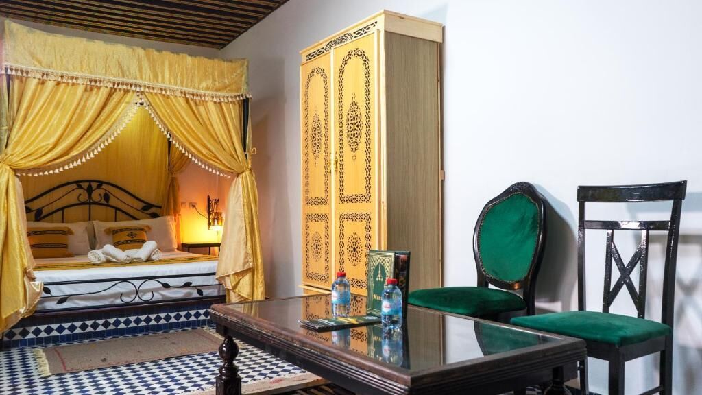 Una habitación con una cama, una silla verde, una mesa con botellas de agua y un mueble decorativo.