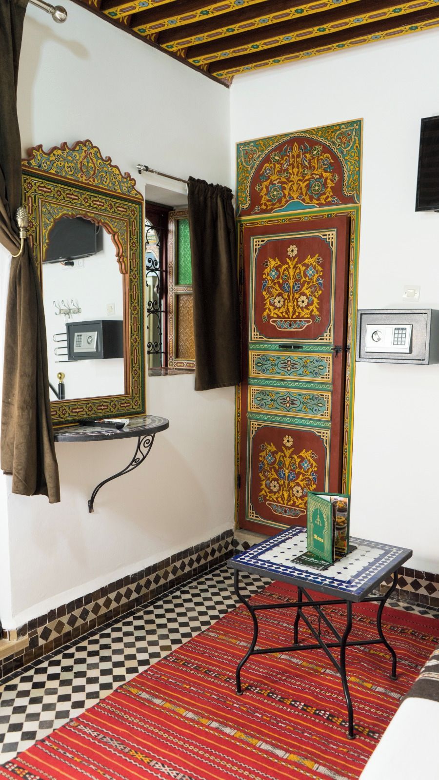 Um quarto com chão xadrez, um espelho com moldura ornamentada, uma porta com desenhos detalhados e uma pequena mesa com um tabuleiro de xadrez.