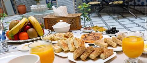 Una mesa con desayuno, mostrando una variedad de alimentos, como frutas, pasteles y bebidas, sobre un piso de baldosas y una decoración tradicional.