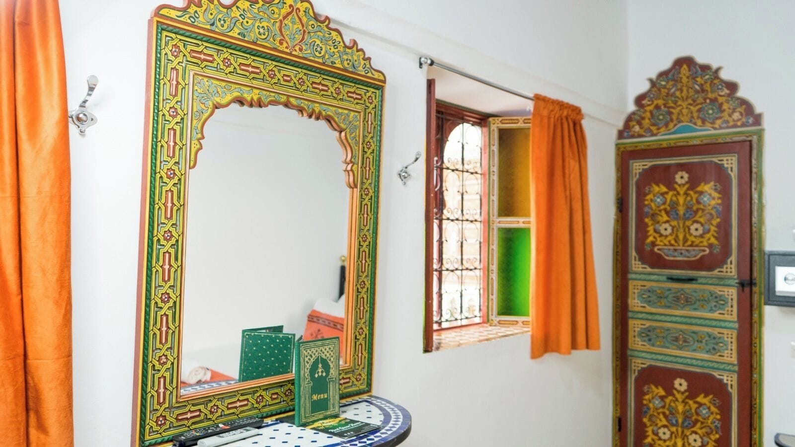 A room with a large ornate mirror, a window with orange curtains, and a door with intricate designs.