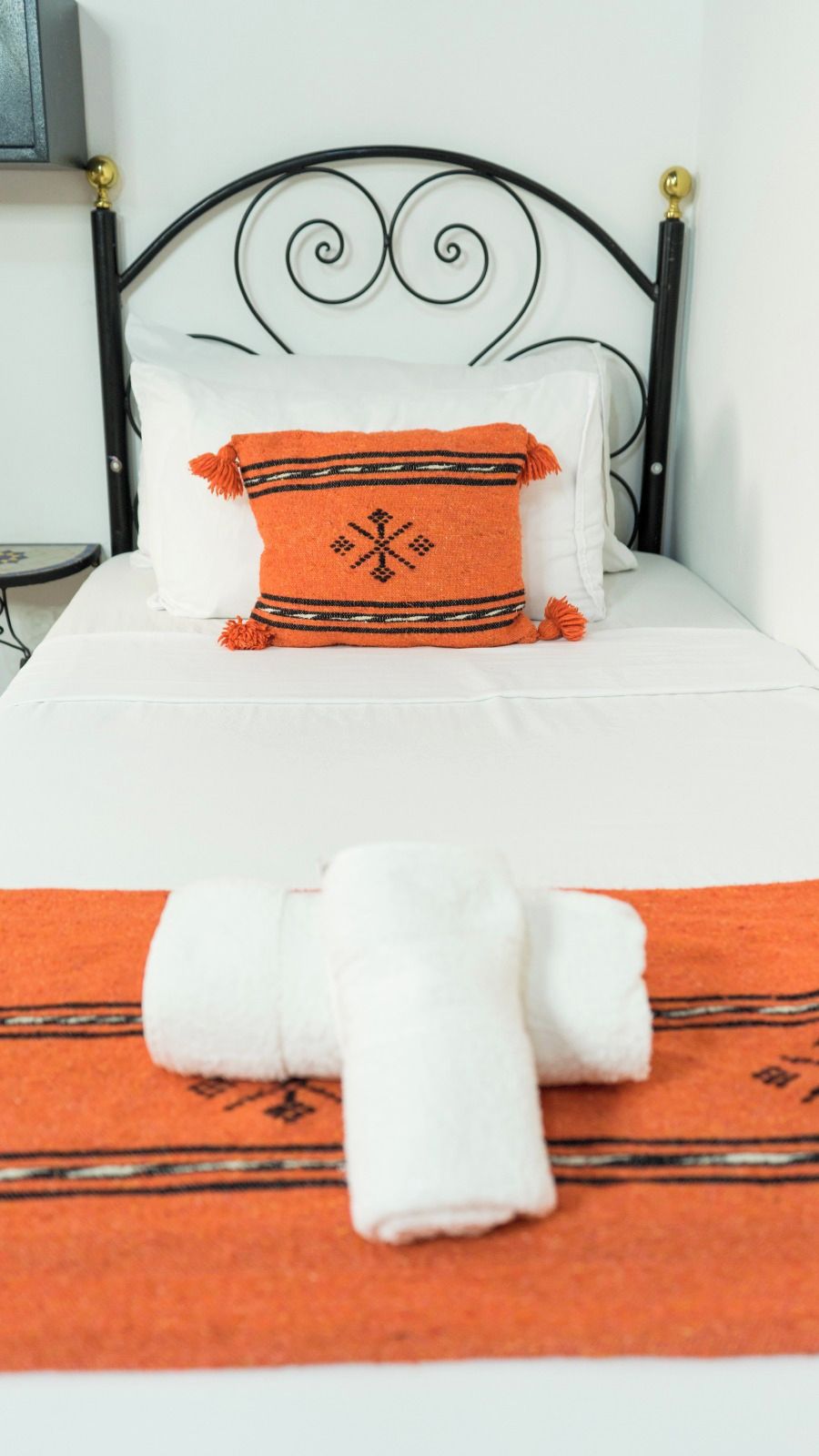 Een bed met een oranje deken en kussens, een witte spreideken, en een opgerolde witte handdoek op een bijpassende oranje deken.