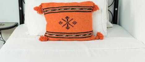 Een bed met een oranje deken en kussens, een witte spreideken, en een opgerolde witte handdoek op een bijpassende oranje deken.
