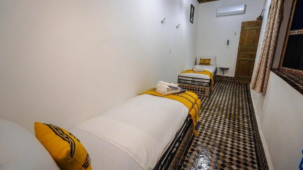 A narrow hallway with two beds, each with a yellow and white bedspread. The floor has a black and white checkered pattern.