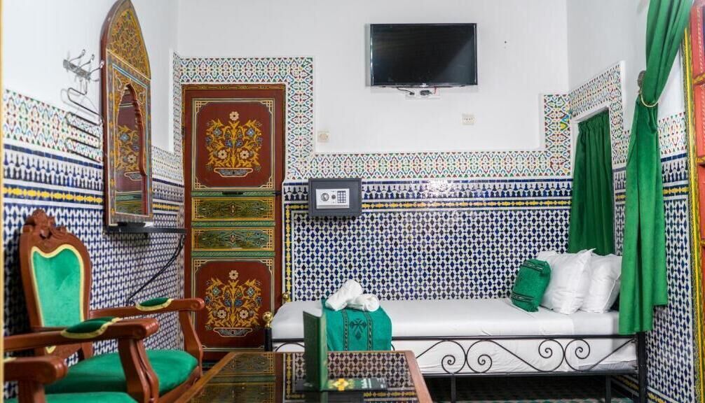 A room with patterned tile walls, a television mounted on the wall, a green sofa, and a wooden chair with a green cushion.