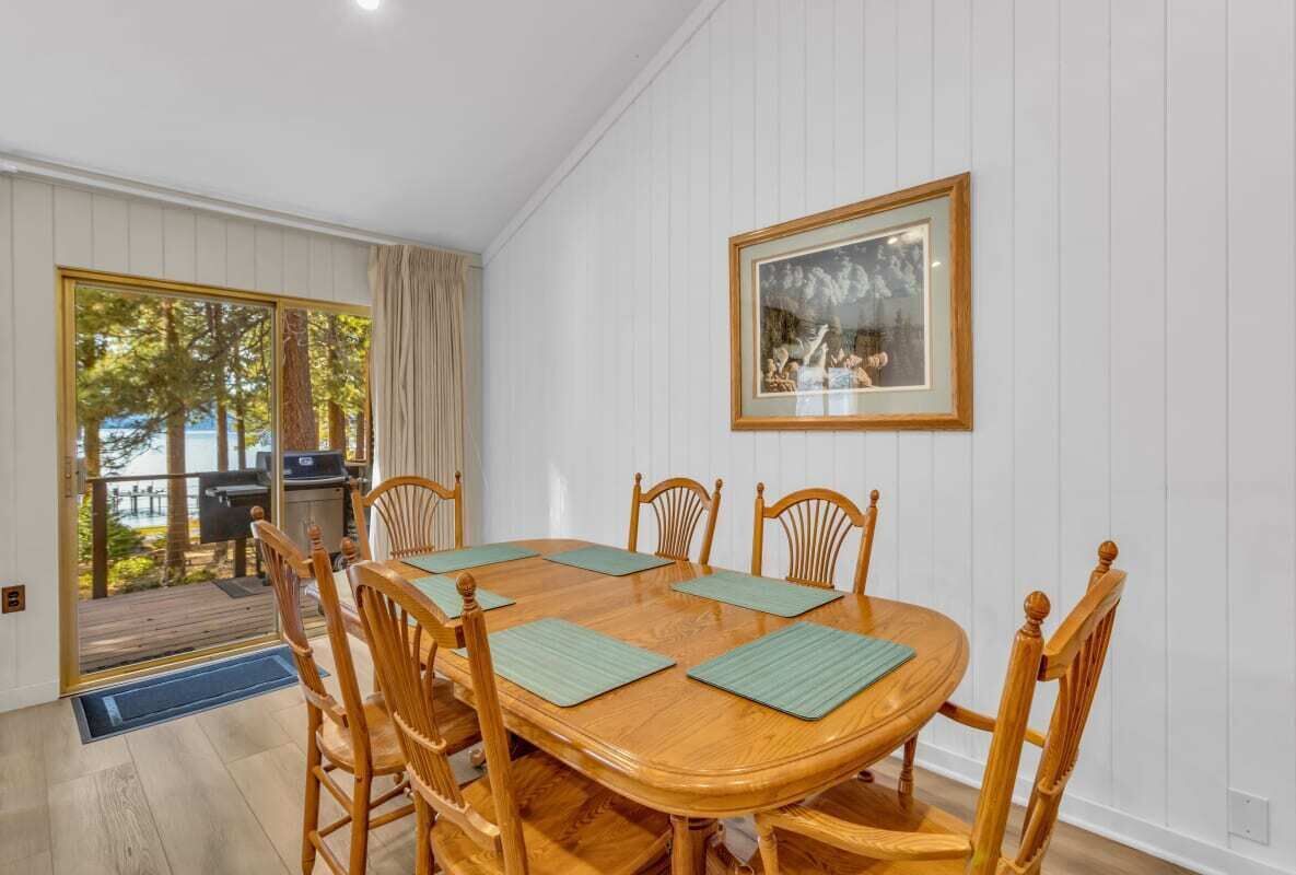 A dining area with a wooden table and chairs, a framed picture on the wall, and a view of the outdoors through a glass door.