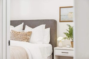 Una cama bien tendida con ropa de cama blanca y beige, un cabecero gris y una mesita de noche con una lámpara y una pequeña planta en maceta.