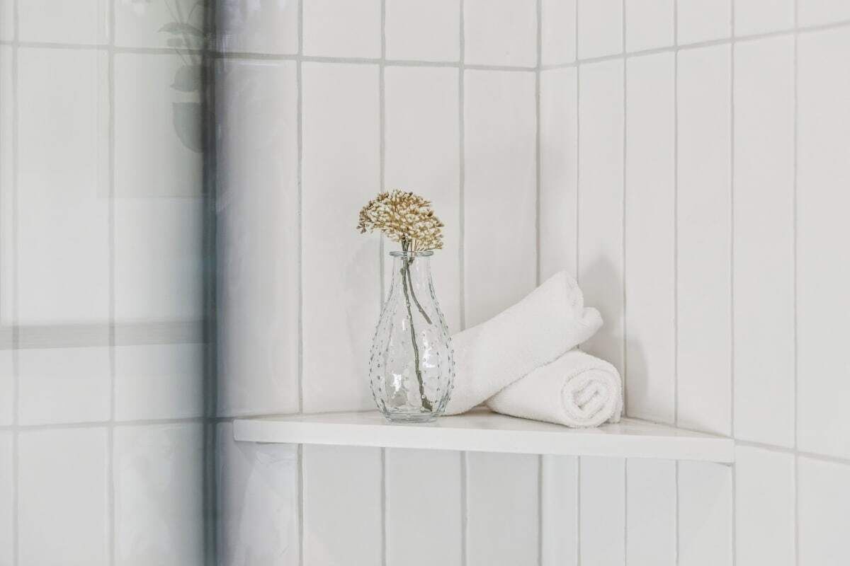 Ein Badezimmer mit weißen Fliesen, einer Glasvase mit Trockenblumen und einem aufgerollten weißen Handtuch auf einem Regal.