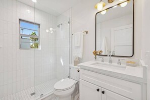 Un baño moderno con ducha de azulejos blancos, inodoro, lavamanos con espejo y ventana con vistas a palmeras.