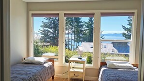 Ein Schlafzimmer mit zwei Betten, einem Schreibtisch und großen Fenstern mit Blick auf Bäume und ein Gebäude.