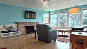 Ein Wohnzimmer mit Kamin aus Stein, einem Flachbildfernseher, einem grauen Sessel, einem runden Holztisch und großen Fenstern mit Blick auf Bäume und den Himmel.