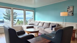 Ein modernes Wohnzimmer mit einer Eckcouch, zwei Sesseln, einem Couchtisch und einer Stehlampe. Große Fenster bieten einen Blick ins Freie.