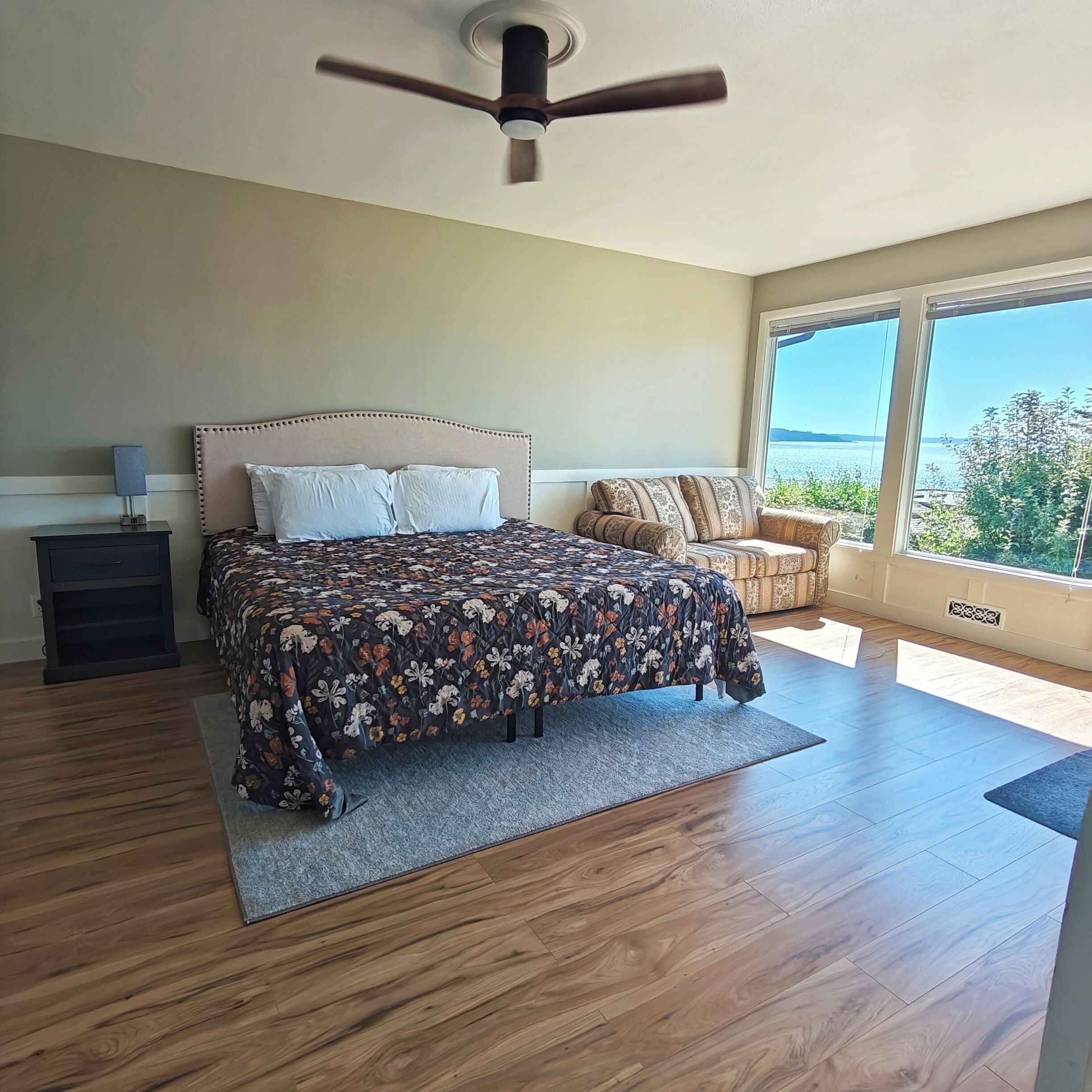 A bedroom with a bed, a nightstand, a ceiling fan, and a view of the sea.