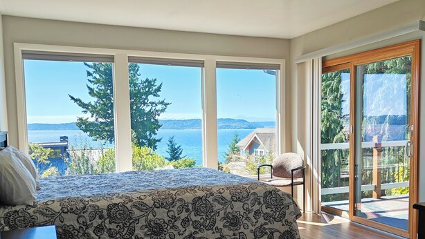 Ein Schlafzimmer mit einem großen Bett, einem Schreibtisch und einem Stuhl. Große Fenster bieten Blick auf das Meer und Bäume.