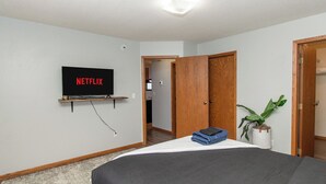 Una camera da letto con un letto, una TV a muro che mostra 'NETFLIX', una pianta e una porta che dà su un'altra stanza.