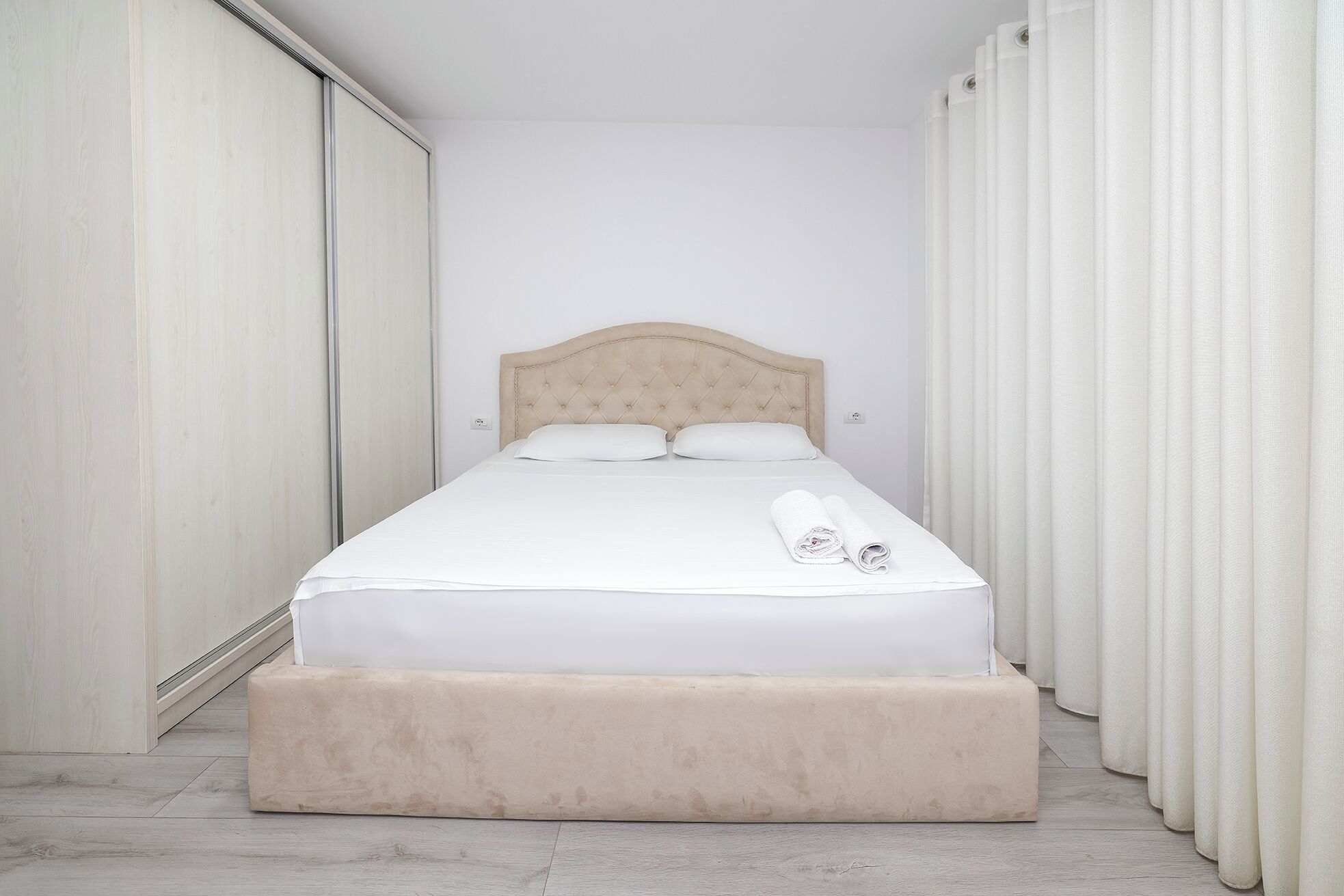 A neatly made bed with a headboard, a rolled towel, and a small ottoman.