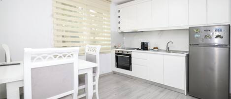 A modern kitchen with white cabinets, a stainless steel refrigerator, a built-in oven, and a window with blinds.