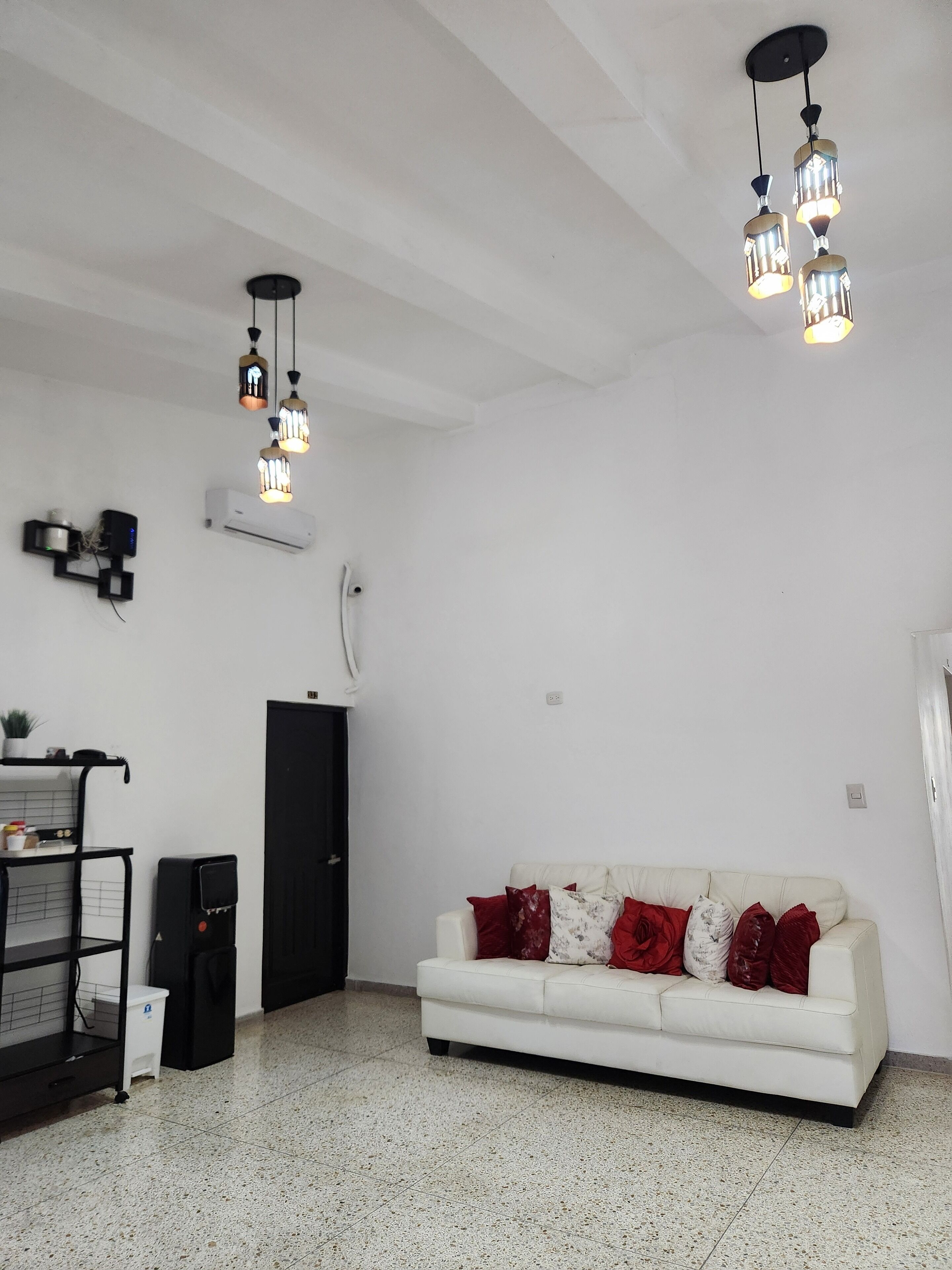 Un salon avec un canapé blanc, des coussins rouges et blancs, un meuble noir, une petite table et un luminaire au plafond.