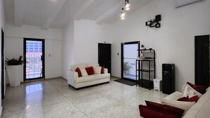 A spacious living area with a white sofa, red cushions, and a coffee table. There are two hanging light fixtures and a black speaker on the wall. - Hotel Feliz Estancia (La Romana)
