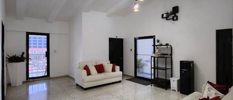 A spacious living area with a white sofa, red cushions, and a coffee table. There are two hanging light fixtures and a black speaker on the wall.