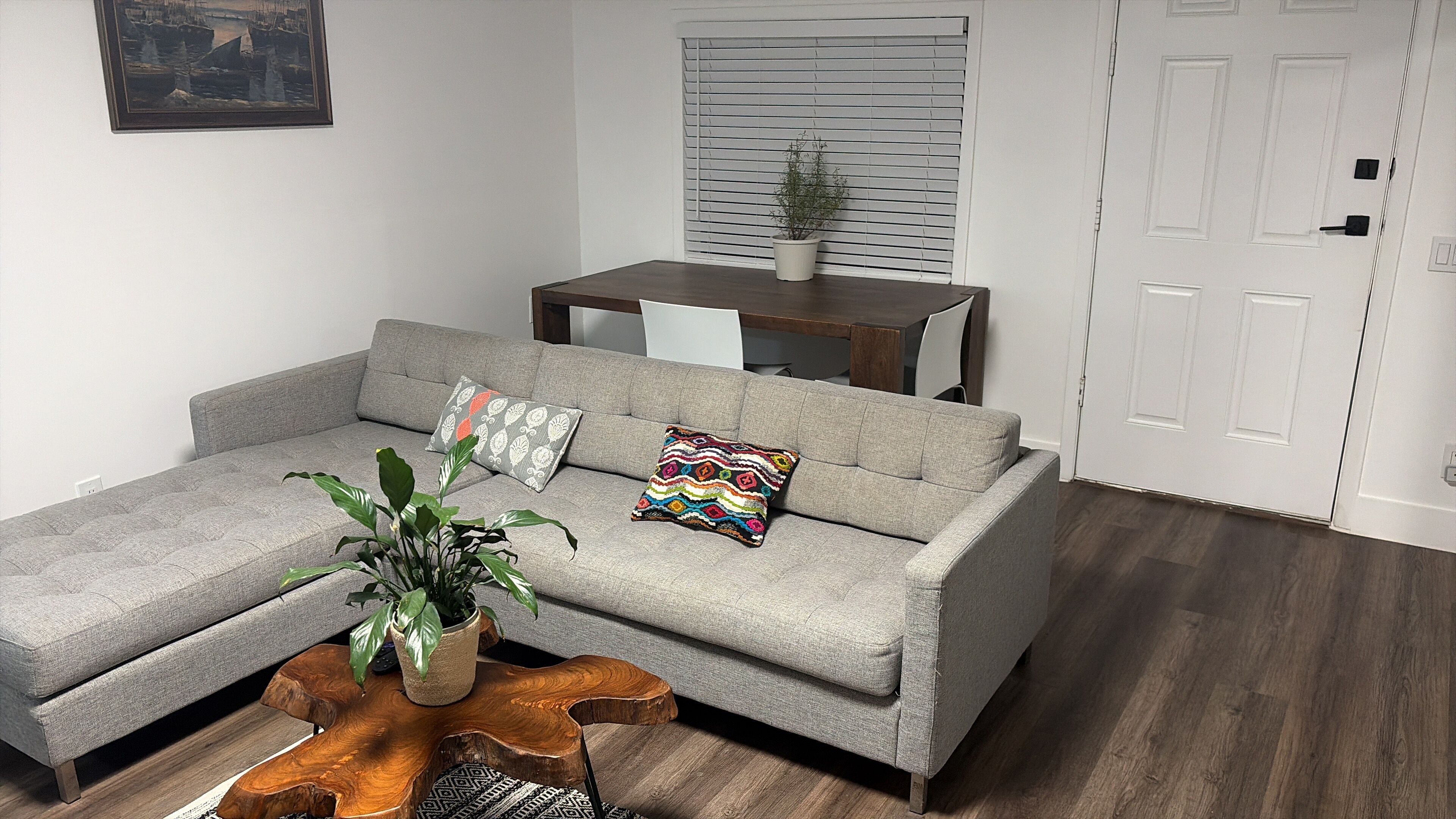 A grey sofa with two patterned cushions, a wooden coffee table with a plant, a dining table with chairs, and a white door.