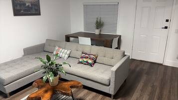 A grey sofa with two patterned cushions, a wooden coffee table with a plant, a dining table with chairs, and a white door.