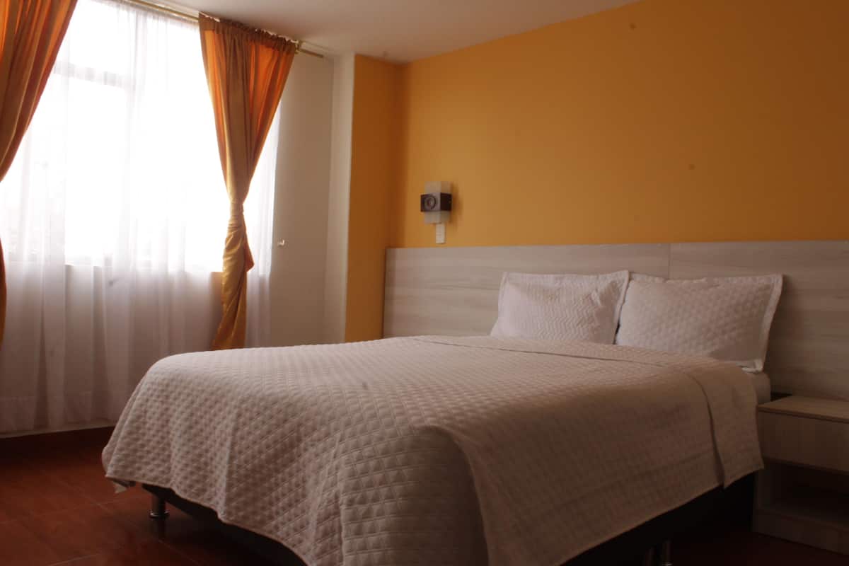 A hotel room with a bed, curtains, a wall-mounted device, and a nightstand.