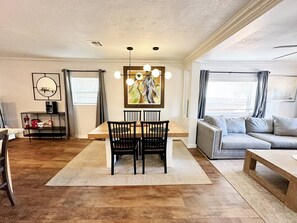Ein modernes Wohnzimmer mit Essbereich, ausgestattet mit einem Holztisch, schwarzen Stühlen, einem grauen Sofa und einem Gemälde an der Wand.