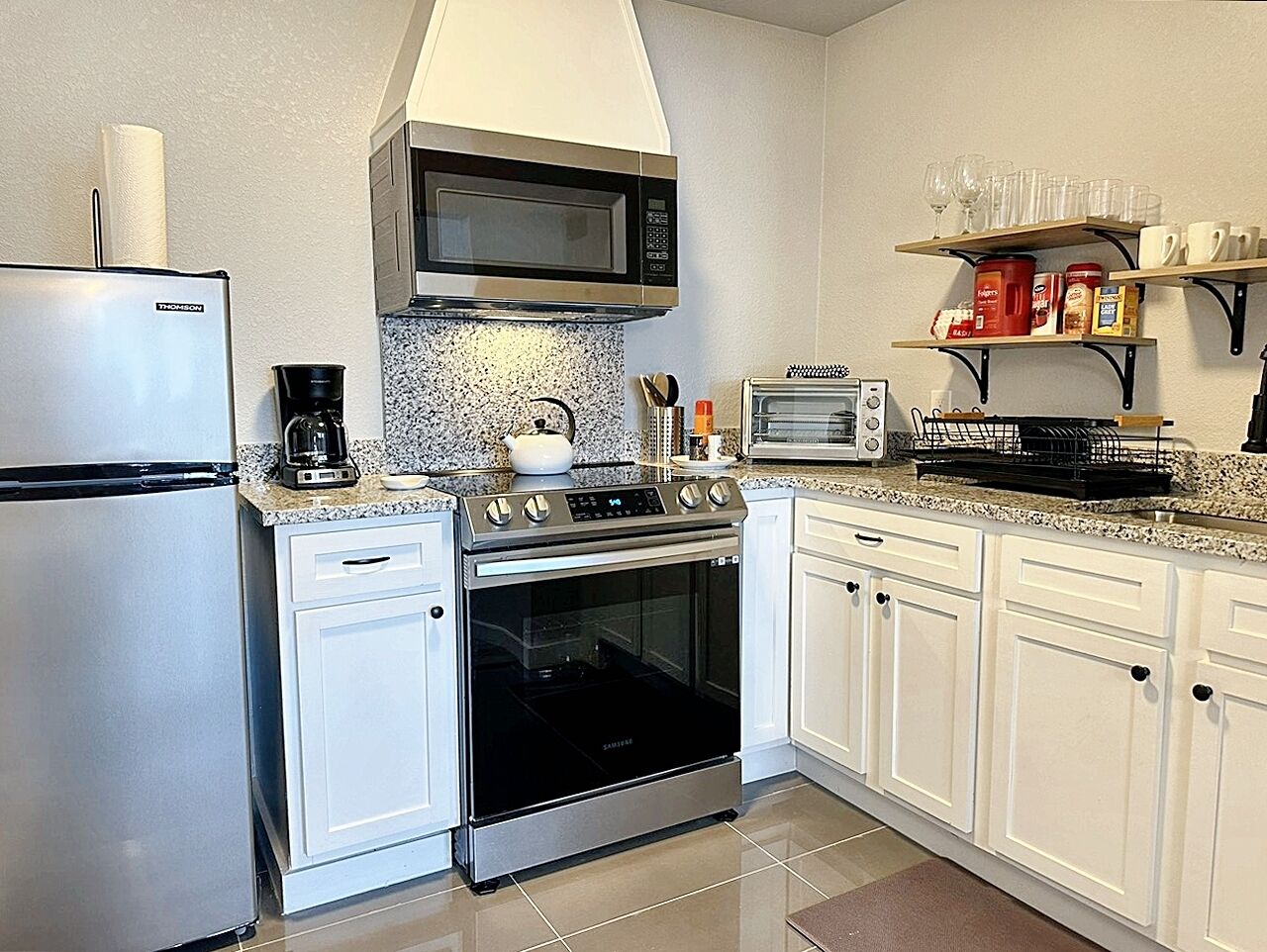 Eine kompakte Küche mit Edelstahlkühlschrank, Mikrowelle, Backofen und Toaster. Weiße Schränke und eine Arbeitsplatte aus Granit.
