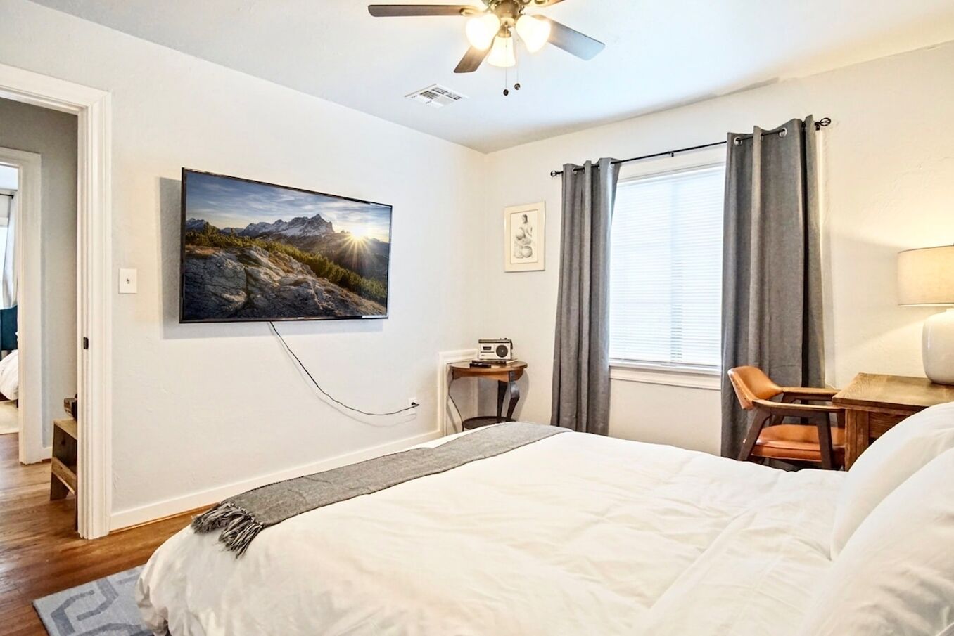 A hotel room with a large bed, a desk with a chair, a ceiling fan, a window with curtains, and a painting on the wall.