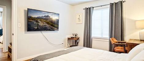 A hotel room with a large bed, a desk with a chair, a ceiling fan, a window with curtains, and a painting on the wall.